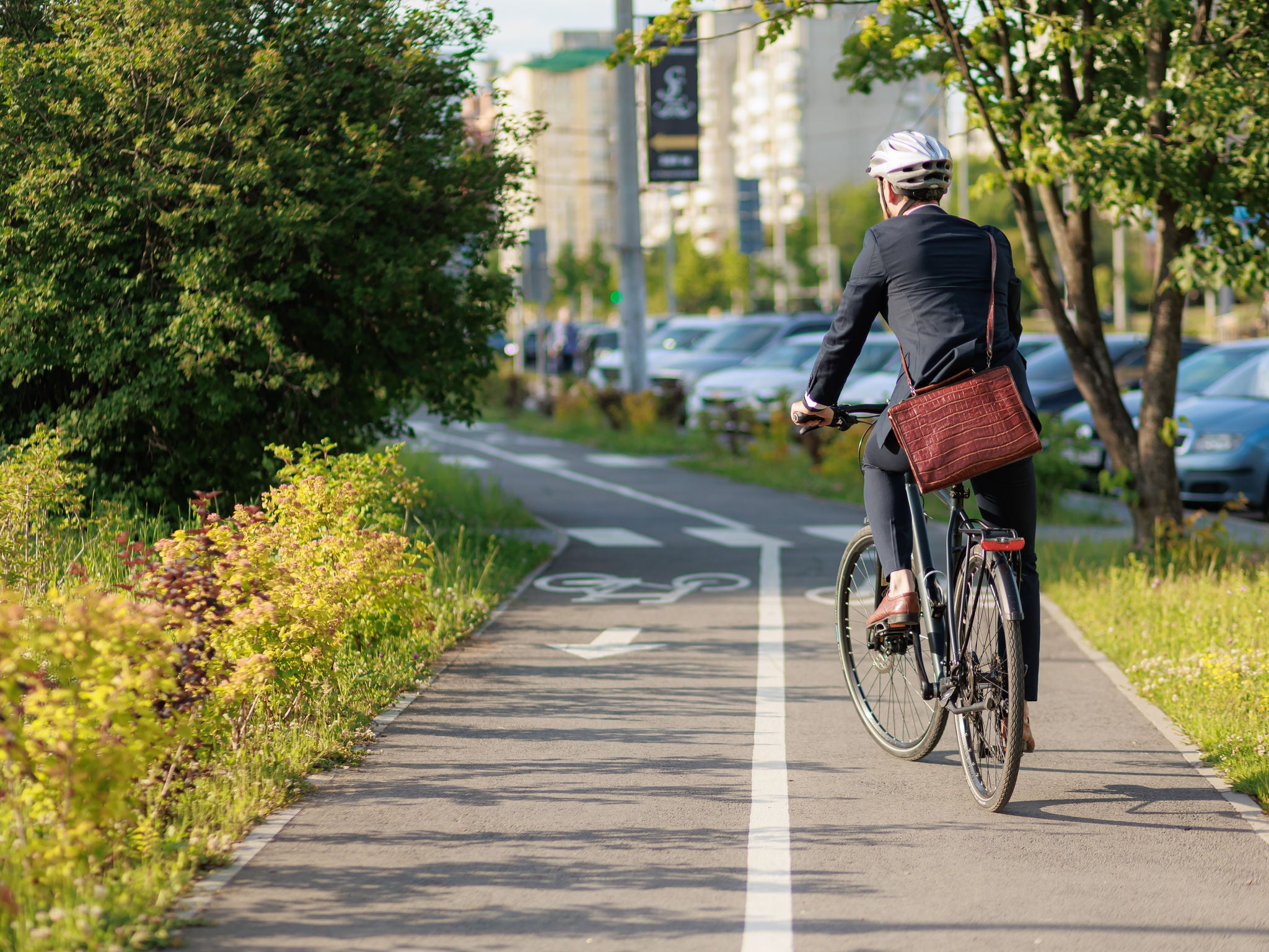 Stylish business man in helmet and suit cycling on bike path in sunny day. Back view of male employee with brown leather briefcase riding on bicycle in town. Concept of eco transport.
